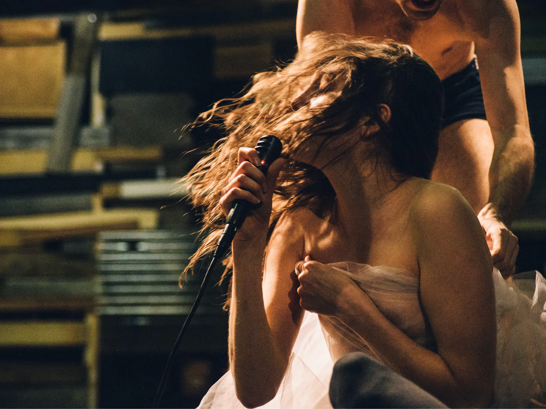 Ženska v gibanju z dolgimi lasmi na odru z mikrofonom, ogrnjena v prosojno blago